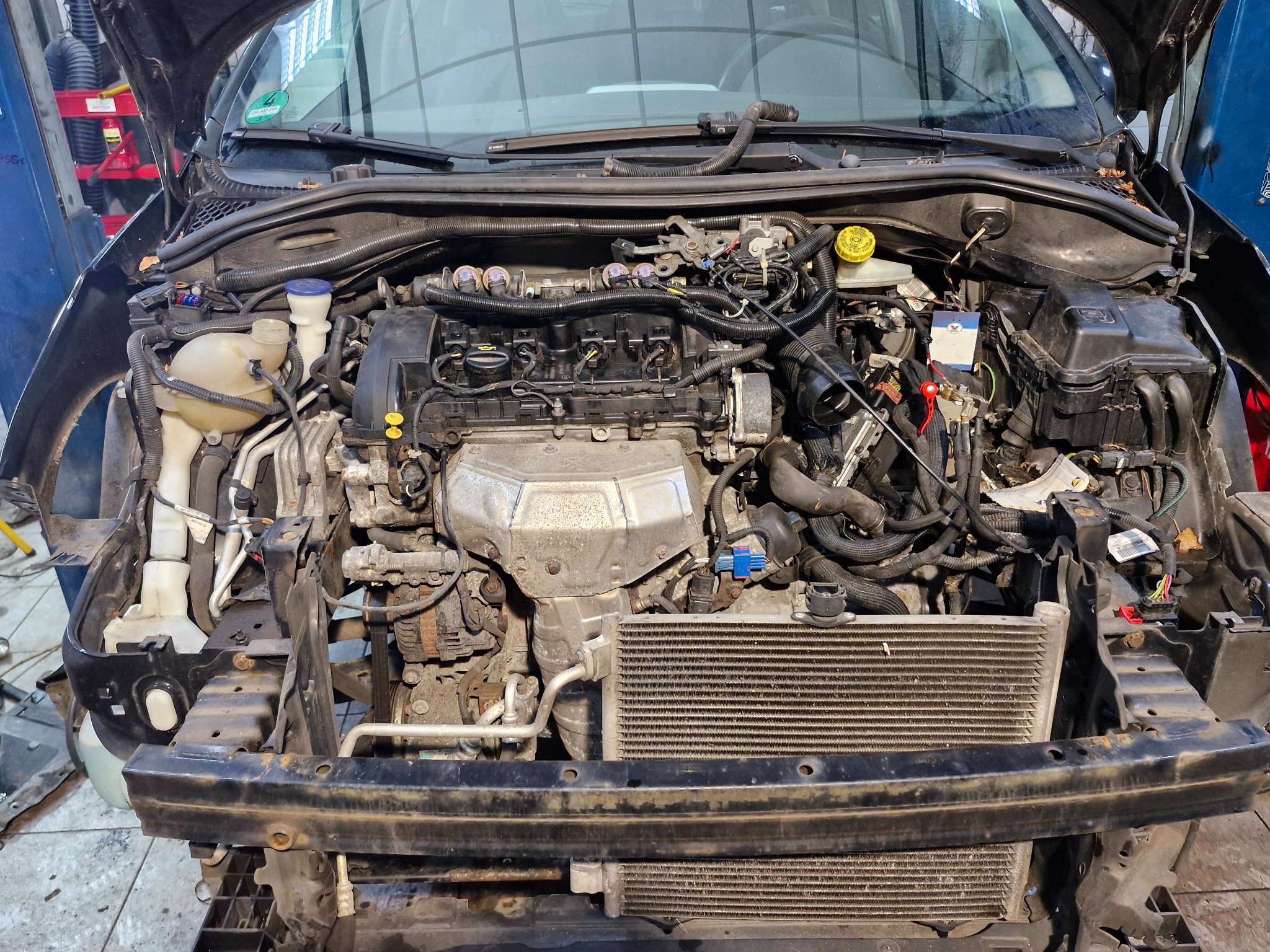 Otwarta maska samochodu z widocznym silnikiem w trakcie naprawy, zdemontowany przedni zderzak, widoczne liczne przewody i elementy mechaniczne.