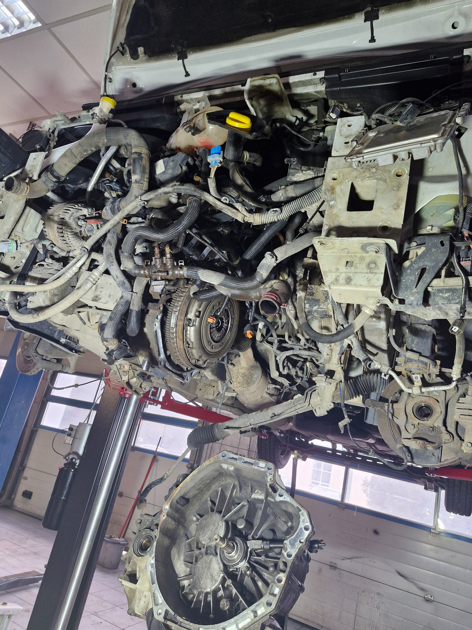 Widok od dołu na podniesiony samochód z odłączoną skrzynią biegów, widoczne liczne przewody i elementy silnika