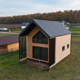 Nowoczesny, drewniany dom z czarnym dachem z blachy na tle jesiennego krajobrazu, widok z lotu ptaka na budynek z dużymi oknami i czarnymi, pionowymi elementami dekoracyjnymi.