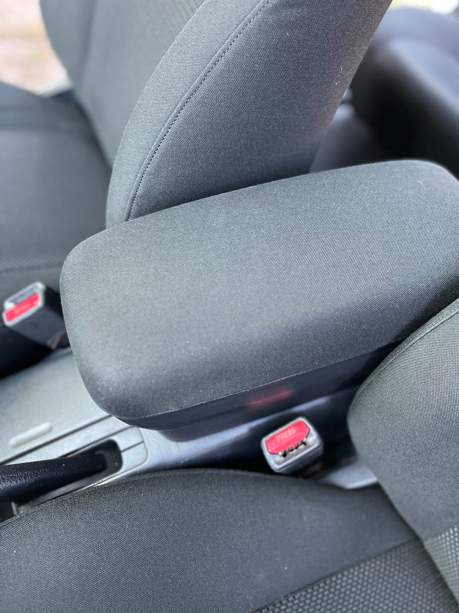 Szczegółowe ujęcie wnętrza samochodu z widocznym podłokietnikiem i fragmentem fotela obitego ciemną tkaniną. Widoczny również pas bezpieczeństwa z czerwonym przyciskiem.