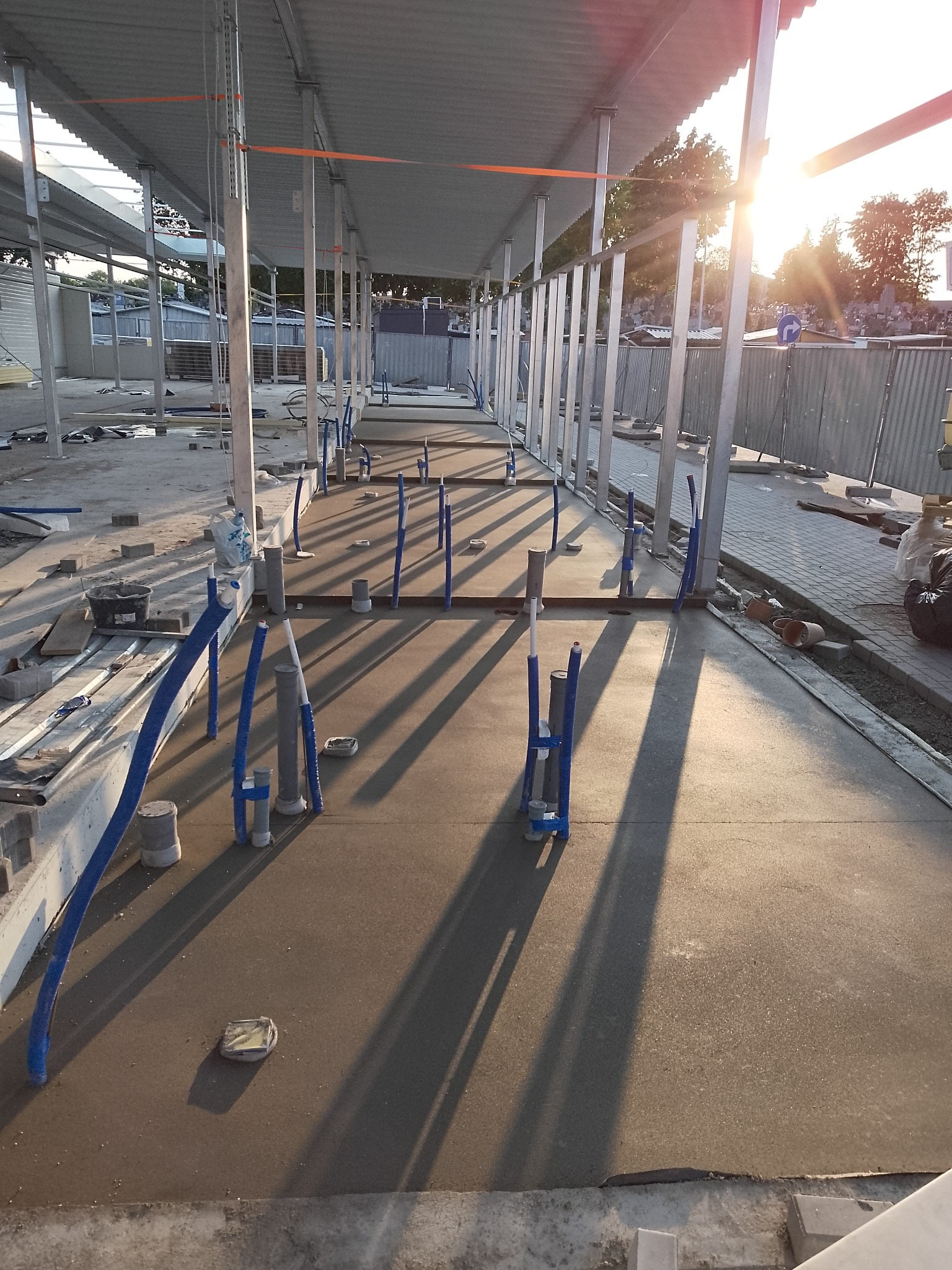 Świeżo wylana, gładka posadzka jastrychowa w hali, z wystającymi rurami instalacji. Długie cienie słupów i rur podkreślają równość powierzchni. Słońce w tle.