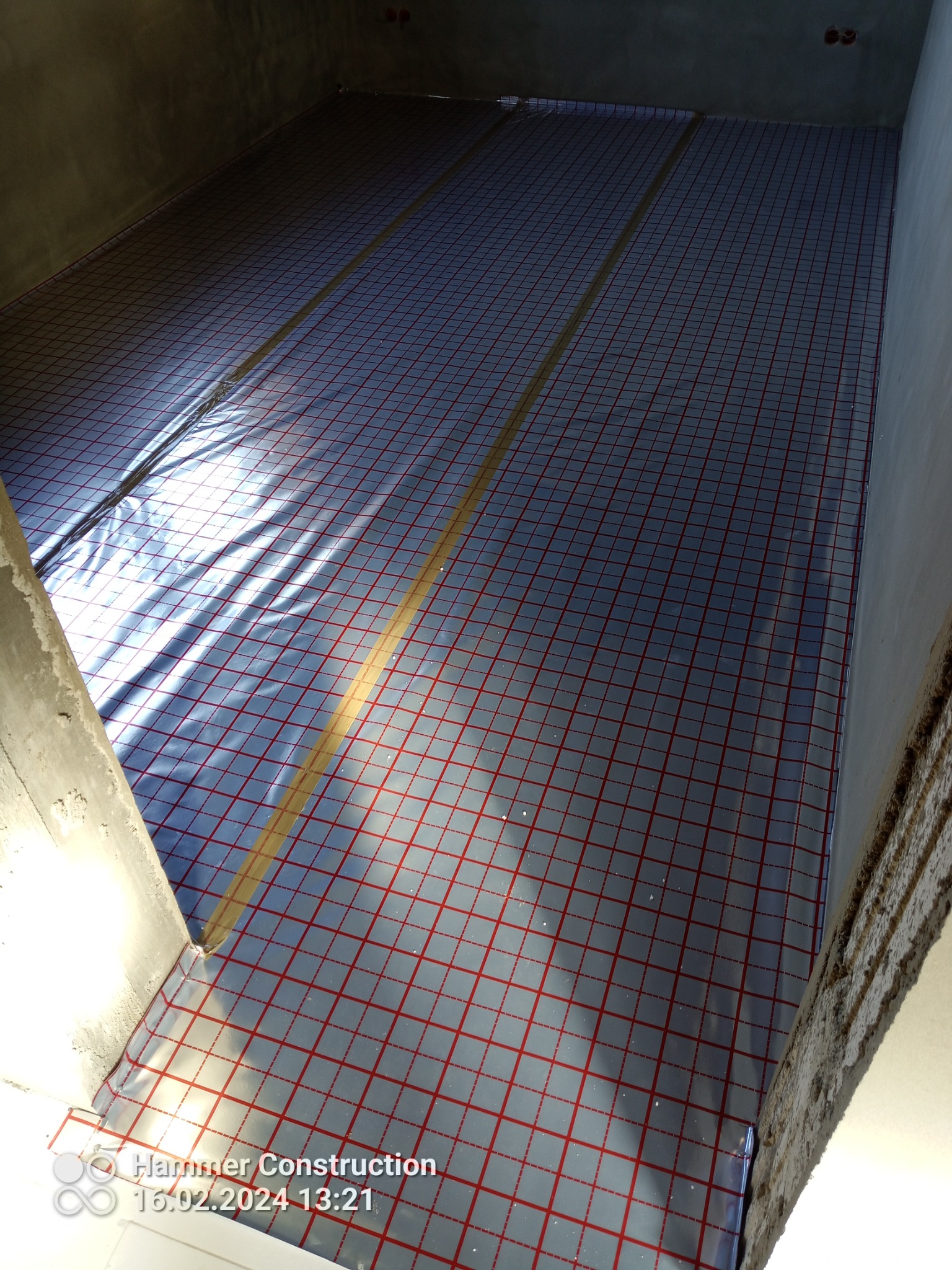 Przygotowanie podłoża pod ogrzewanie podłogowe: folia z siatką w czerwone kwadraty rozłożona na surowej podłodze w pomieszczeniu z betonowymi ścianami, widoczne fragmenty taśmy klejącej.