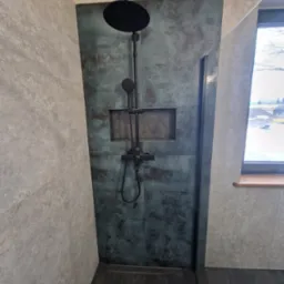 Kabina prysznicowa walk-in z czarną armaturą i płytkami imitującymi beton w odcieniach szarości i turkusu, z półką w ścianie i odpływem liniowym.