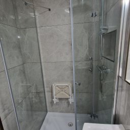 Remonty Karol Jurenko - Szklana kabina prysznicowa z siedziskiem na ścianie wyłożonej szarymi płytkami imitującymi marmur, widoczna bateria i deszczownica.