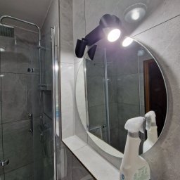 Remonty Karol Jurenko - Nowoczesna łazienka z szarą marmurową okładziną, kabiną prysznicową typu walk-in i okrągłym lustrem z oświetleniem punktowym; na blacie stoi butelka środka czyszczącego.