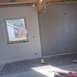 Remonty Karol Jurenko - Wnętrze w trakcie prac wykończeniowych: częściowo pomalowana ściana na szaro, widoczne przygotowania do ogrzewania podłogowego, okno z widokiem na zewnątrz, żółte wiaderko i narzędzia na podłodze...