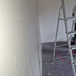 Remonty Karol Jurenko - Świeżo nałożona gładź na ścianie, widoczna aluminiowa drabina i zabezpieczona podłoga przed dalszymi pracami wykończeniowymi.