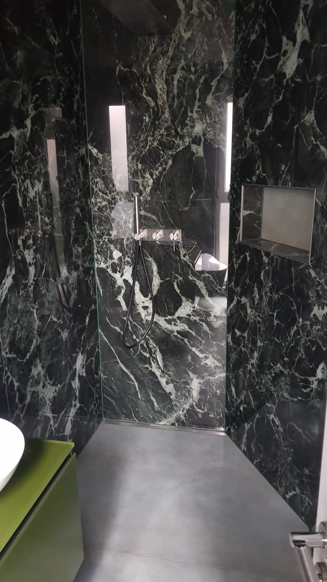 Kabina prysznicowa z czarnym marmurem na ścianach, szara posadzka i zielona szafka pod umywalką.