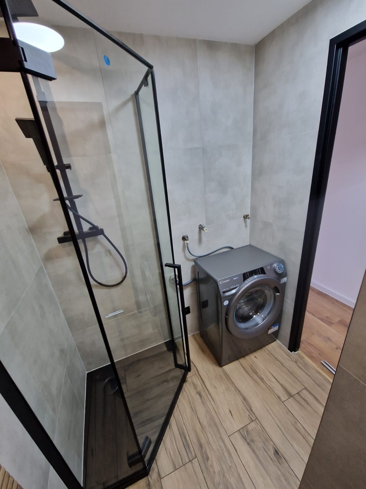 Nowoczesna łazienka z kabiną prysznicową typu walk-in z czarną ramą, szarą pralką Candy i płytkami imitującymi drewno na podłodze i ścianach.