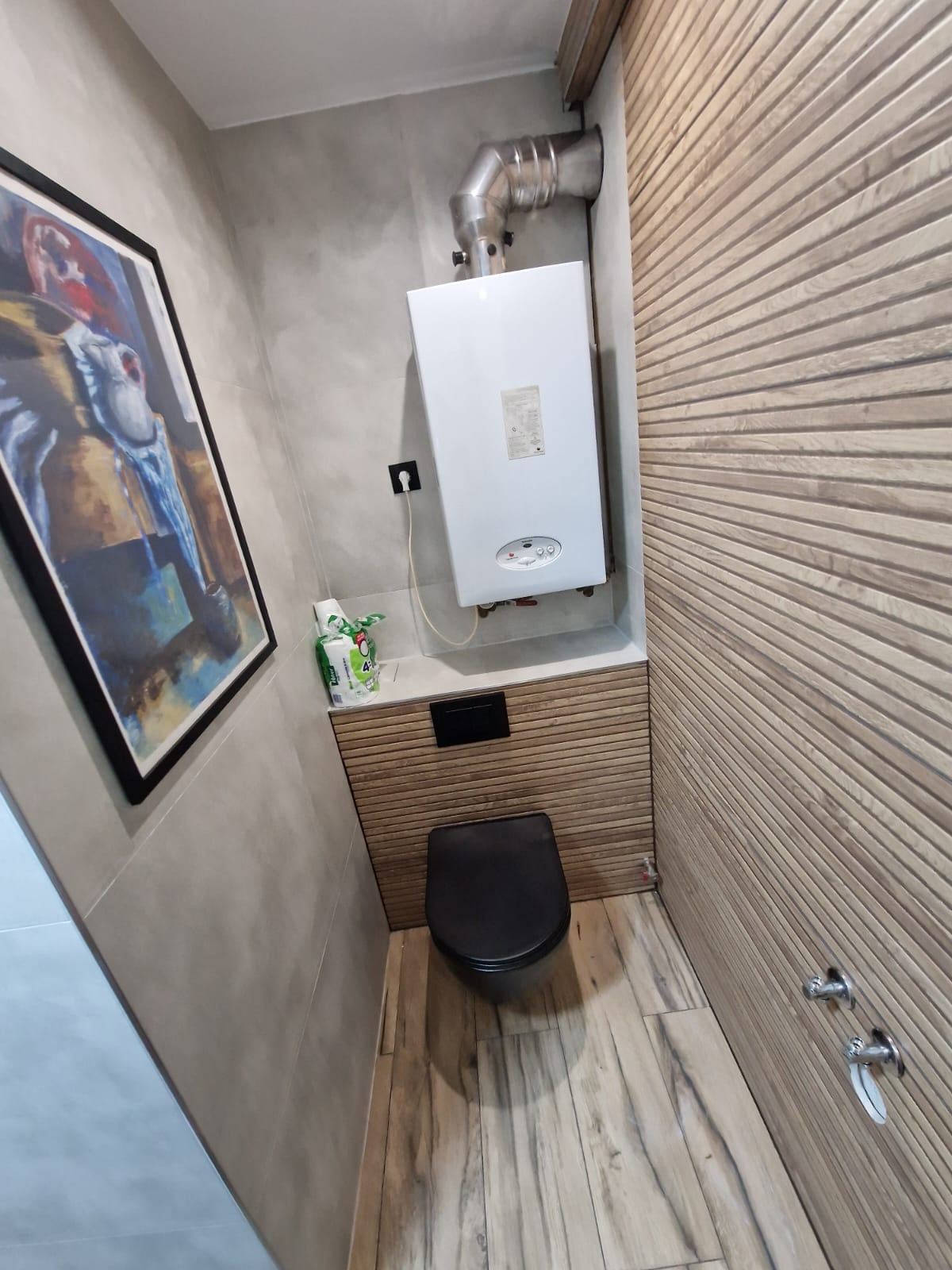 Nowoczesna łazienka z wiszącą toaletą w kolorze czarnym, drewnopodobną okładziną ścian i podłogi, oraz grzejnikiem gazowym. Na ścianie wisi obraz w czarnej ramie.