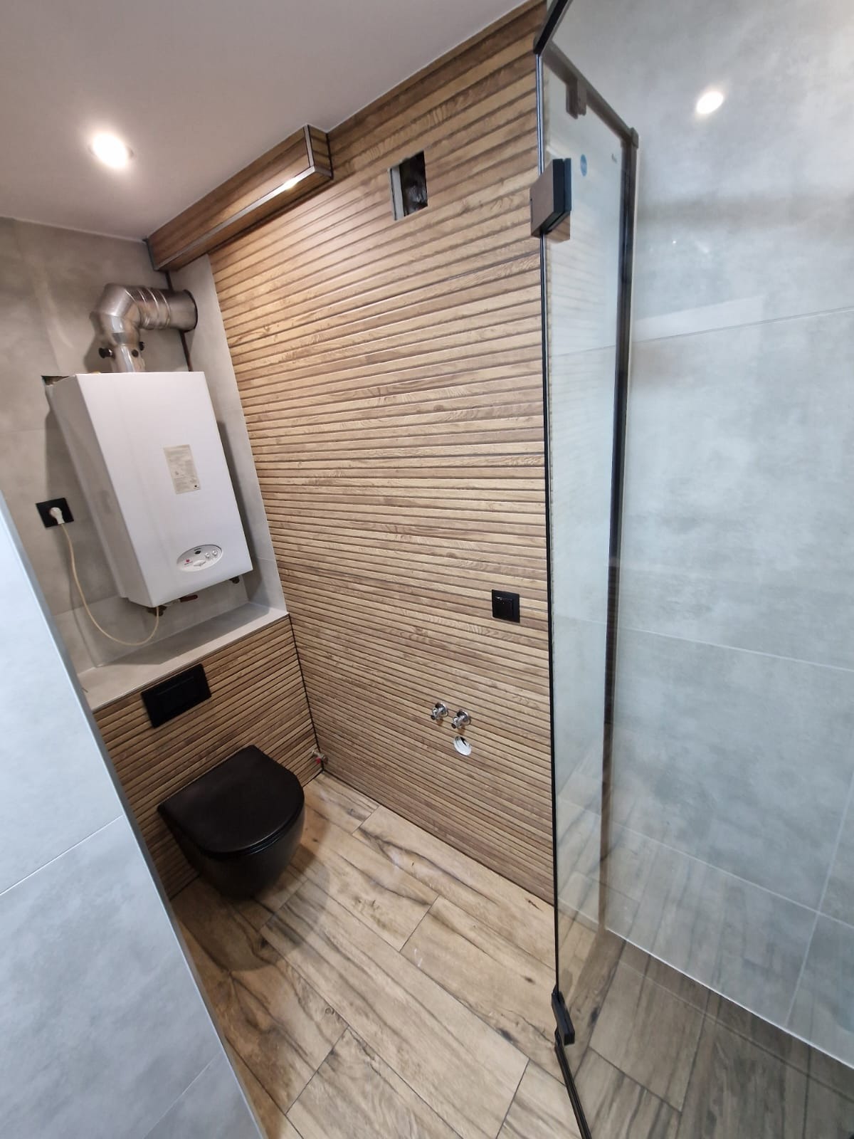 Nowoczesna łazienka z drewnianą ścianą z poziomych paneli, czarną wiszącą toaletą, szklaną kabiną prysznicową i białym piecem grzewczym.