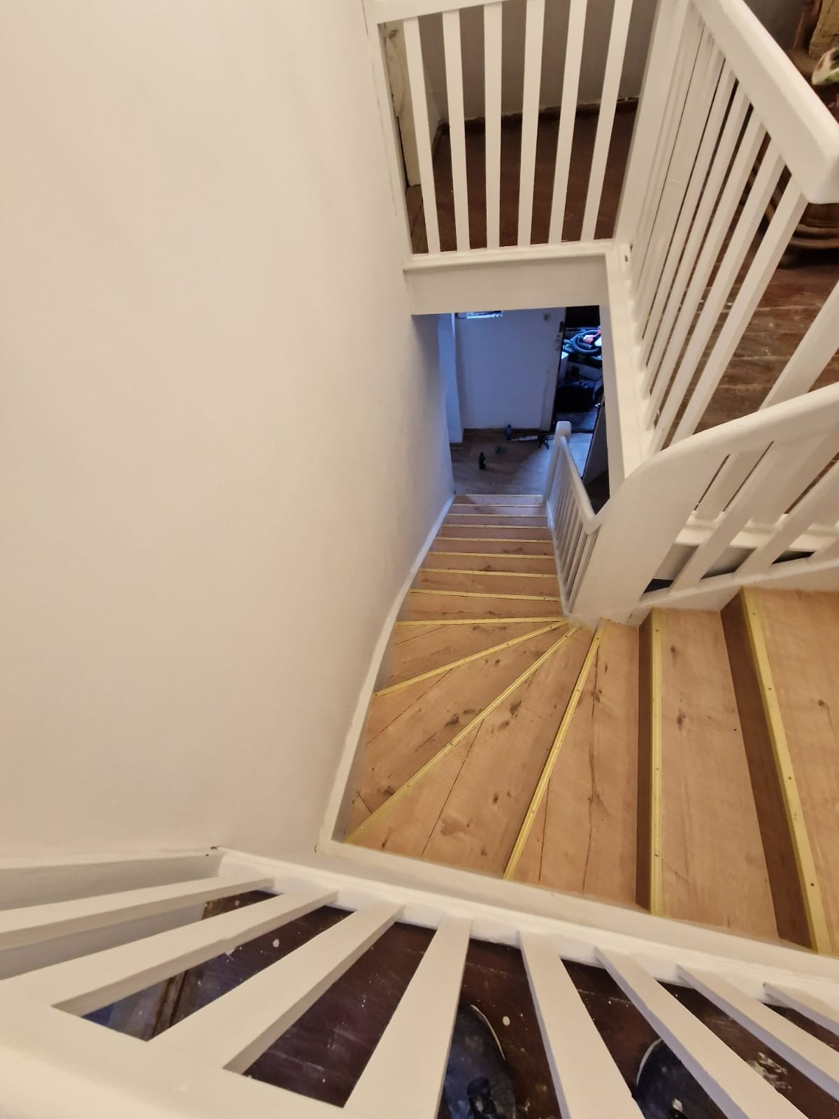 Klatka schodowa w trakcie renowacji: drewniane stopnie zabezpieczone taśmą malarską, białe balustrady i ściany po malowaniu, widok z góry.