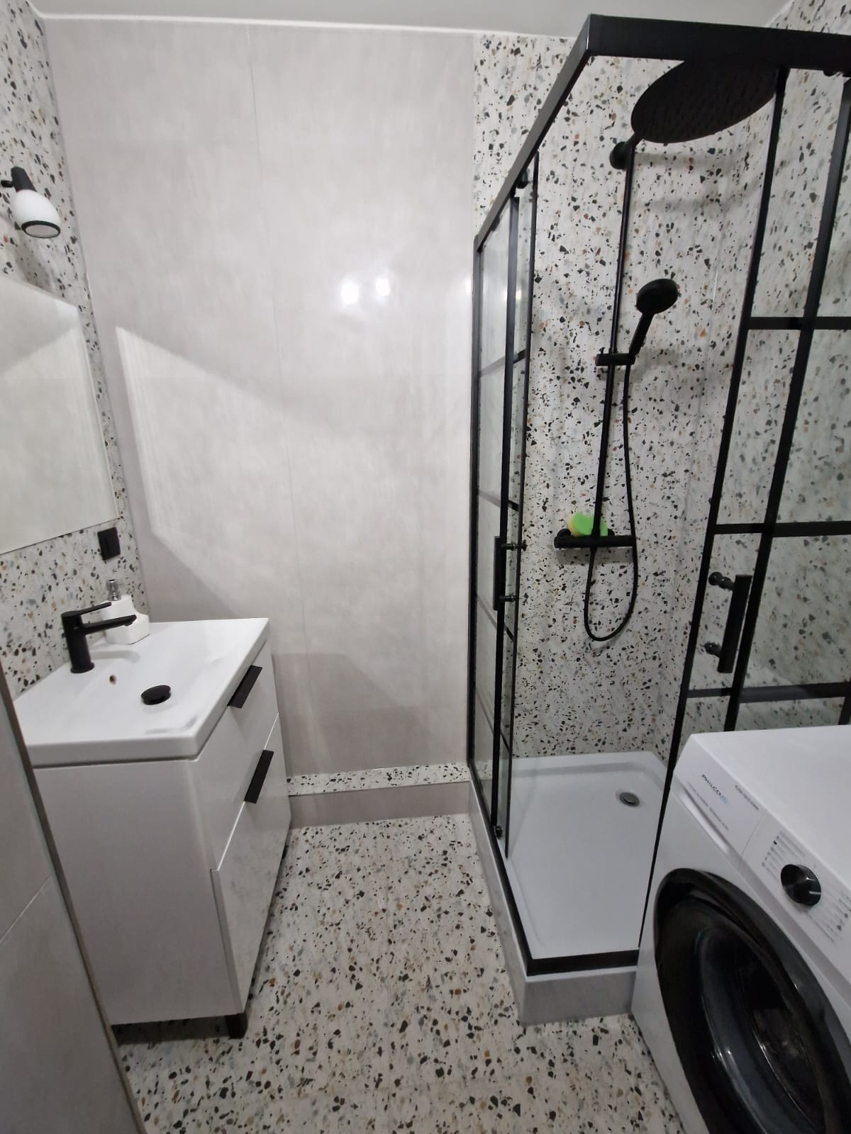 Wykończona łazienka z kabiną prysznicową typu walk-in z czarnymi profilami, umywalką nablatową z szafką w kolorze białym i pralką; ściany i podłoga wykończone płytkami lastryko.