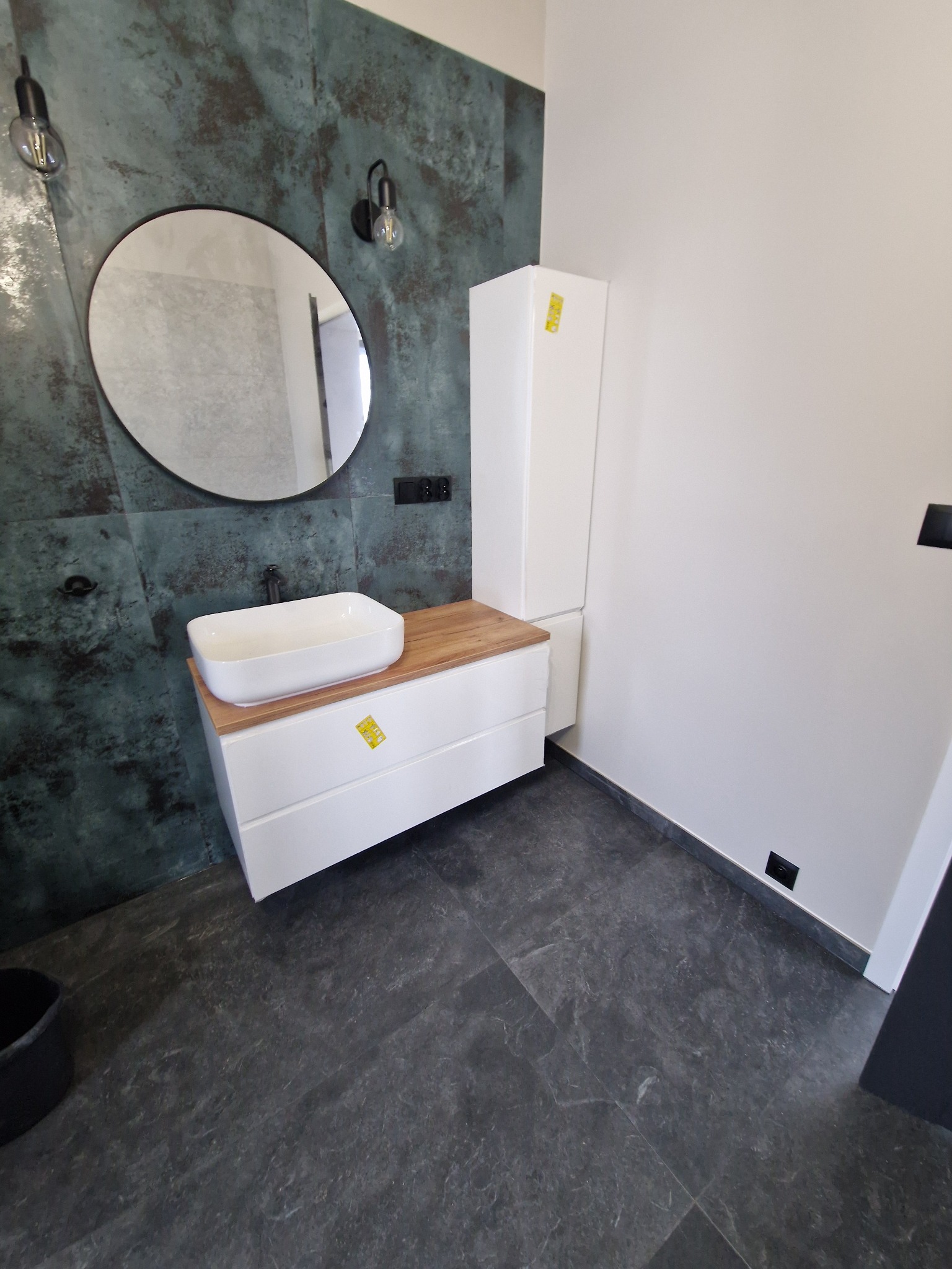 Nowoczesna łazienka z umywalką nablatową, szafką z drewnianym blatem, okrągłym lustrem, dekoracyjną ścianą w odcieniach zieleni i szarości oraz szarą podłogą.