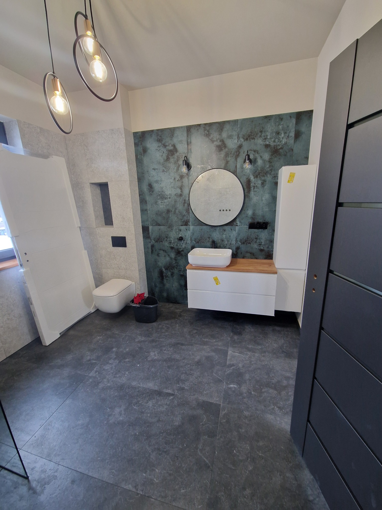 Nowoczesna łazienka w trakcie wykańczania: szara podłoga, umywalka na drewnianym blacie, okrągłe lustro na tle turkusowej ściany, wiszące lampy, biała toaleta i drzwi w folii ochronnej.