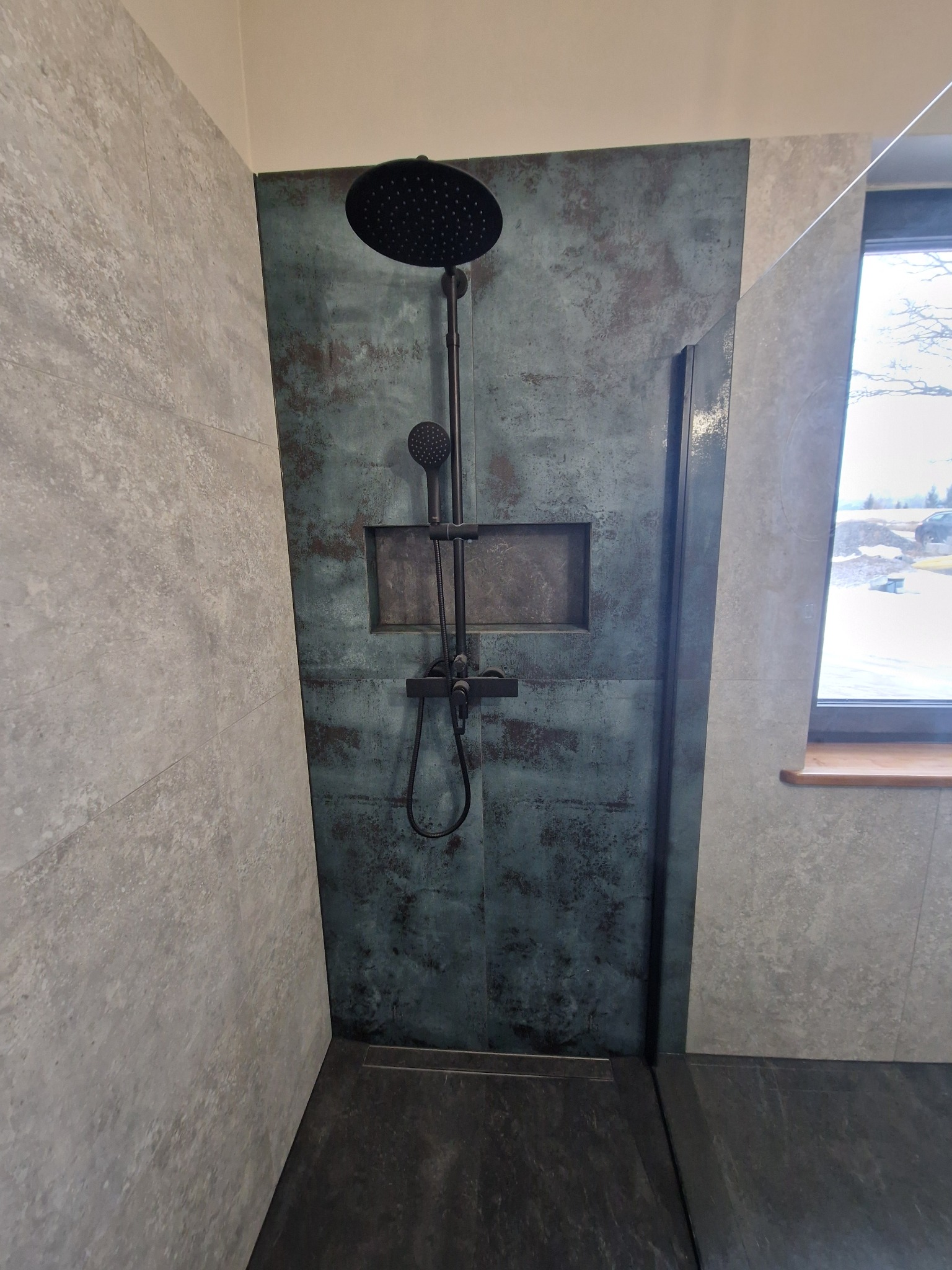 Kabina prysznicowa walk-in z czarną armaturą i płytkami imitującymi beton w odcieniach szarości i turkusu, z półką w ścianie i odpływem liniowym.