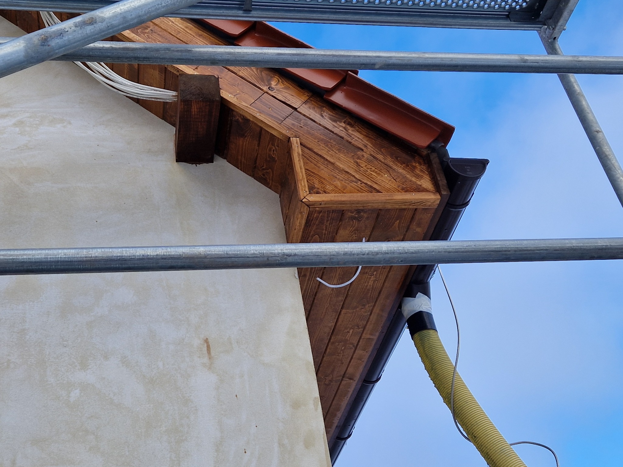 Ocieplony i pomalowany okap dachu z drewnianą podbitką, widok z rusztowania na tle błękitnego nieba.