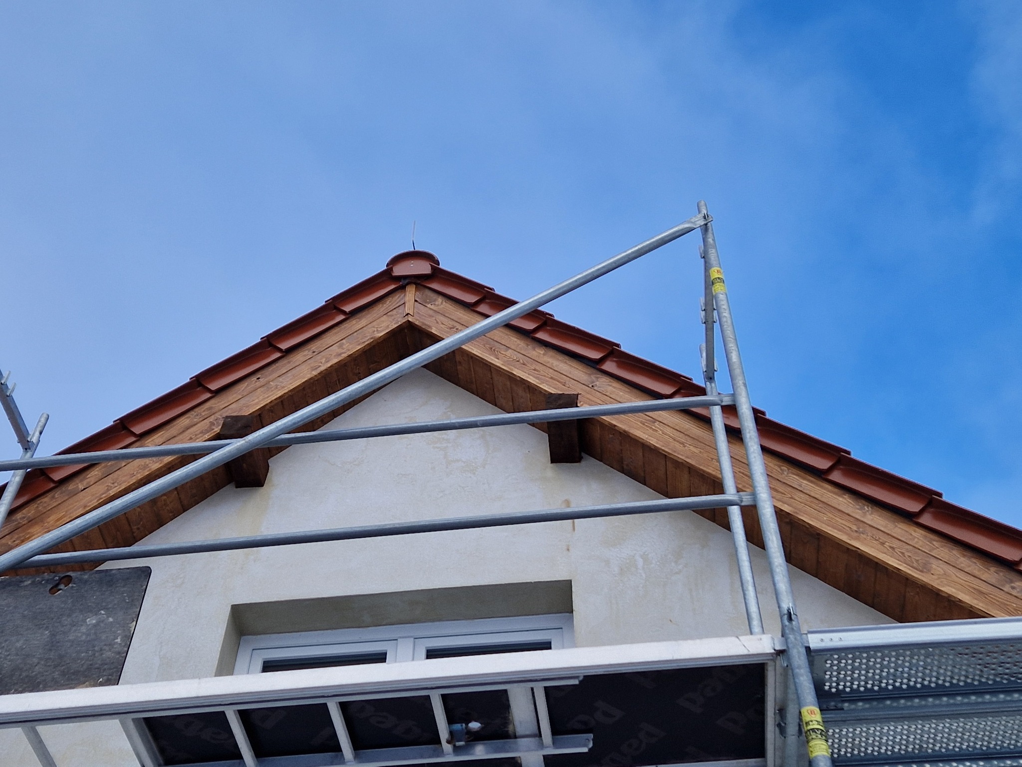 Fragment elewacji domu z drewnianą podbitką i brązową dachówką, widoczny spod rusztowania na tle błękitnego nieba. Na ścianie widoczne ślady po pracach wykończeniowych.