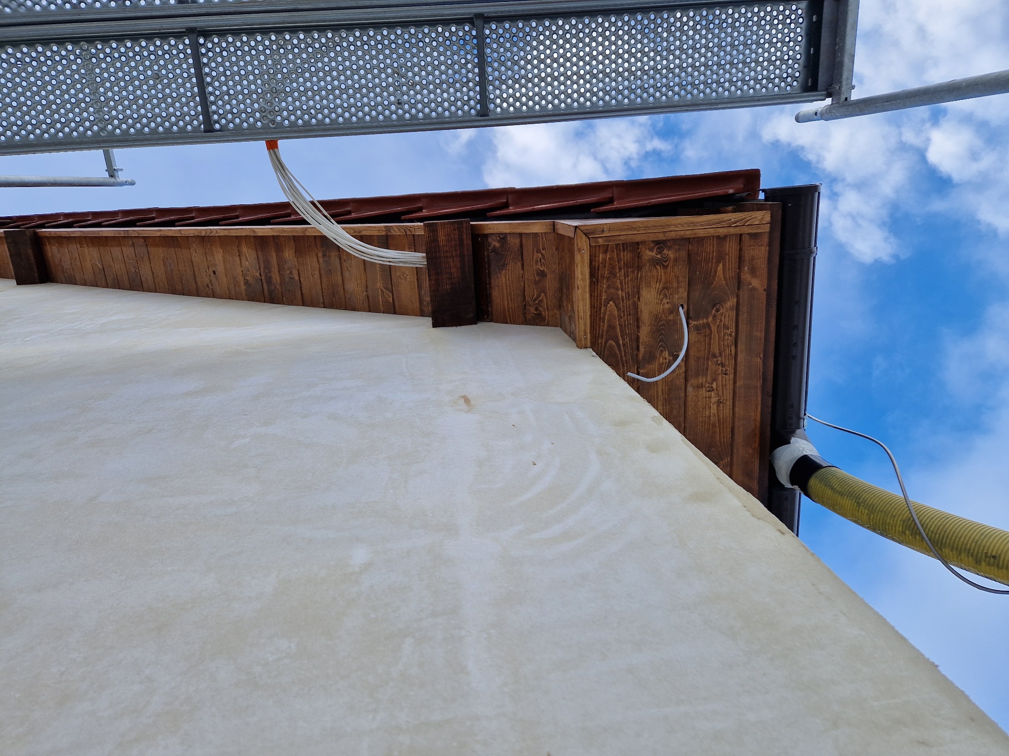 Elewacja budynku z drewnianym wykończeniem pod dachem, widoczne rusztowanie i przewody elektryczne oraz rura wentylacyjna, ujęcie z dołu.