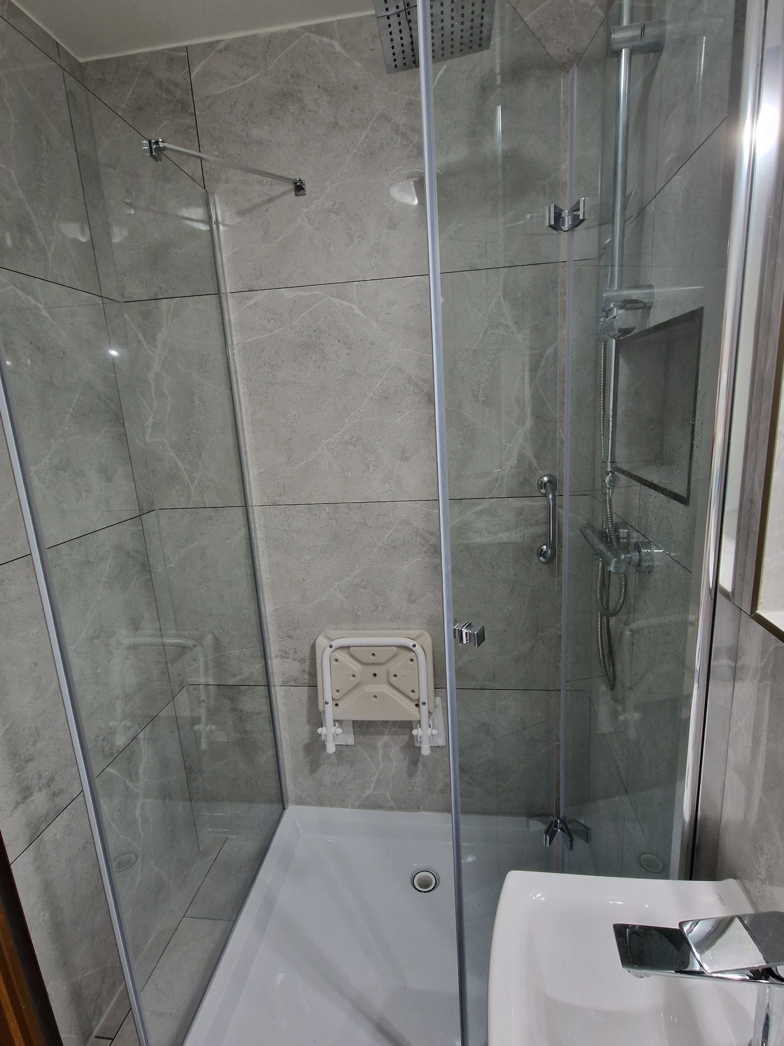 Szklana kabina prysznicowa z siedziskiem na ścianie wyłożonej szarymi płytkami imitującymi marmur, widoczna bateria i deszczownica.