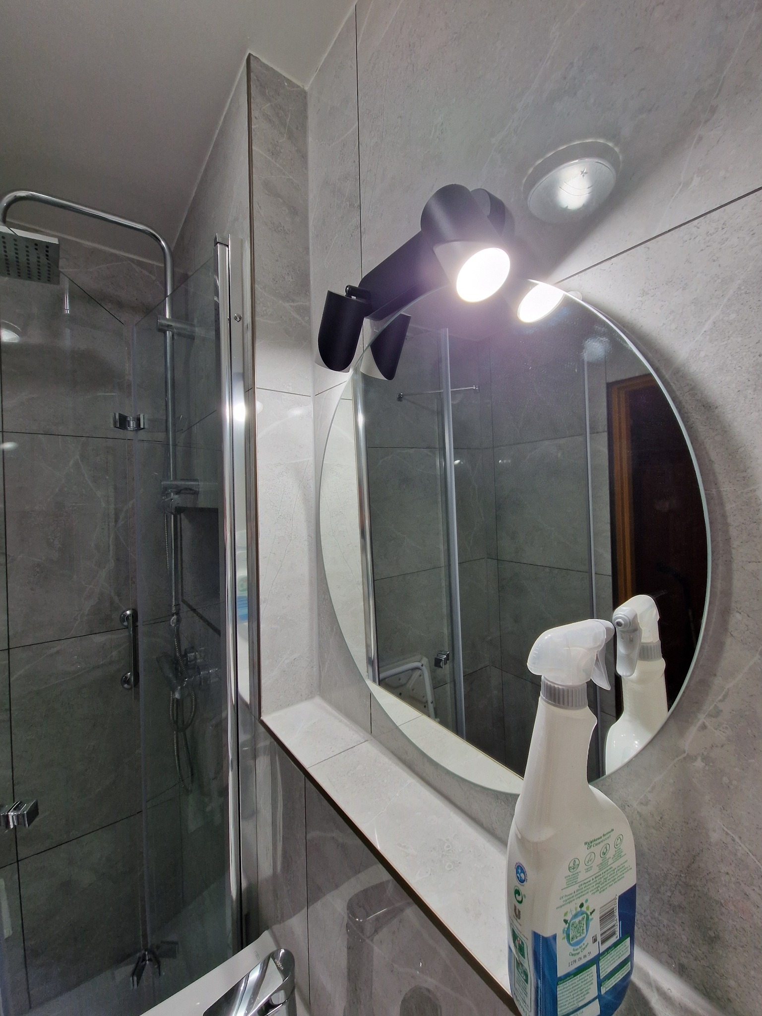 Nowoczesna łazienka z szarą marmurową okładziną, kabiną prysznicową typu walk-in i okrągłym lustrem z oświetleniem punktowym; na blacie stoi butelka środka czyszczącego.