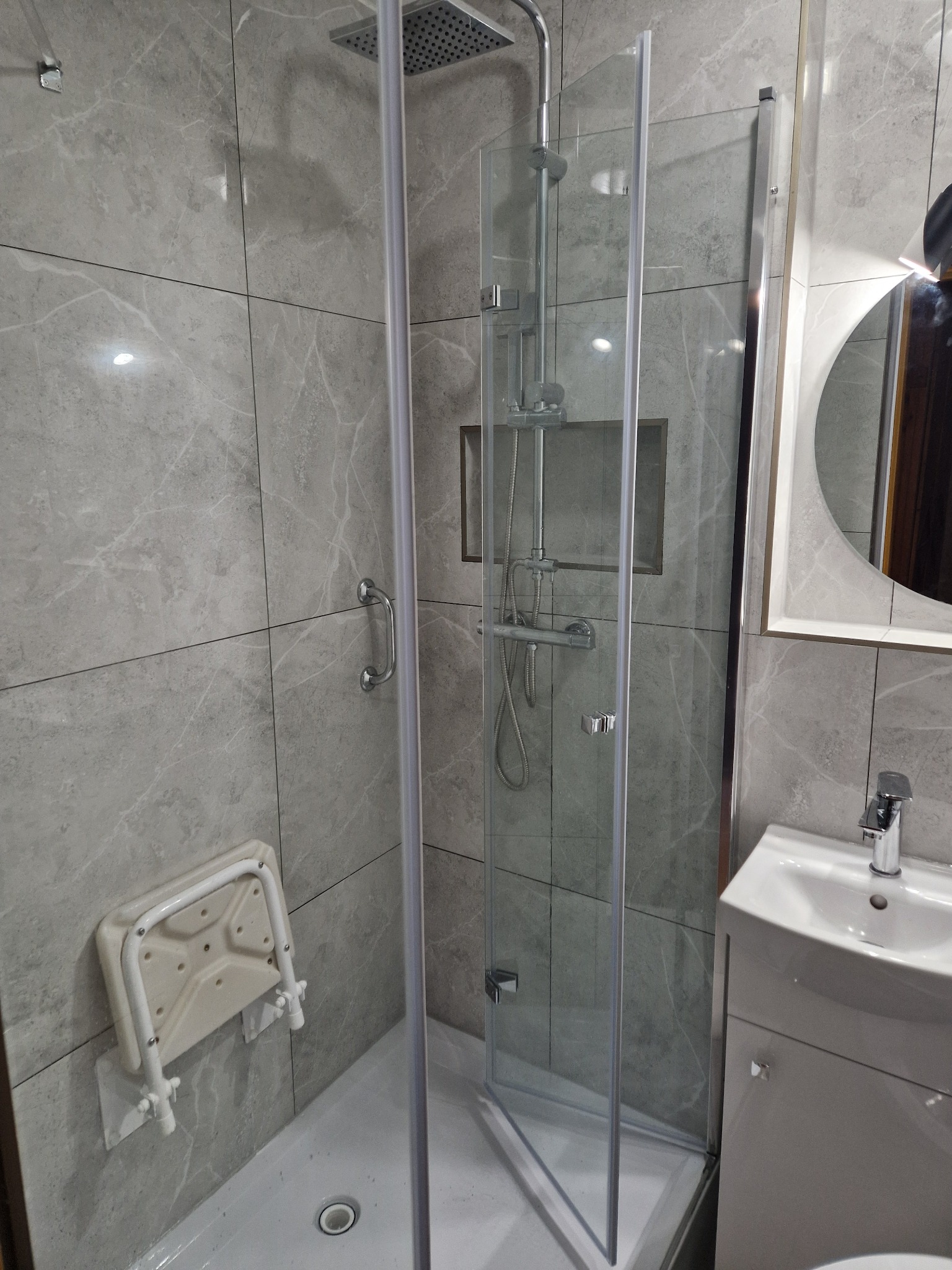 Nowoczesna kabina prysznicowa z szarymi marmurkowymi płytkami, składanym siedziskiem i okrągłym lustrem z podświetleniem LED.