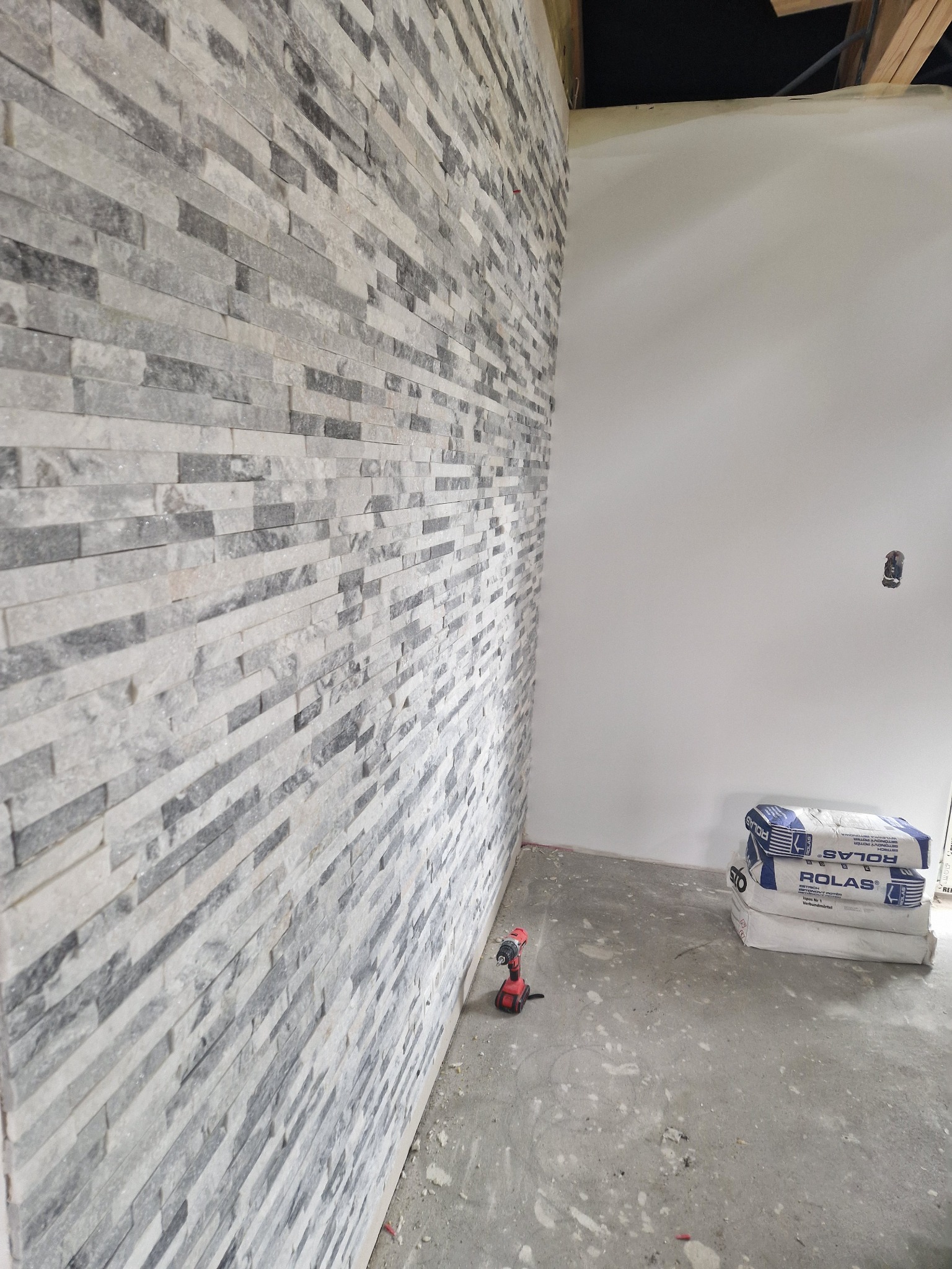Ściana wyłożona szarymi płytkami imitującymi kamień, z widocznym wiertłem akumulatorowym i workami zaprawy klejowej ROLAS na podłodze, w trakcie prac wykończeniowych.