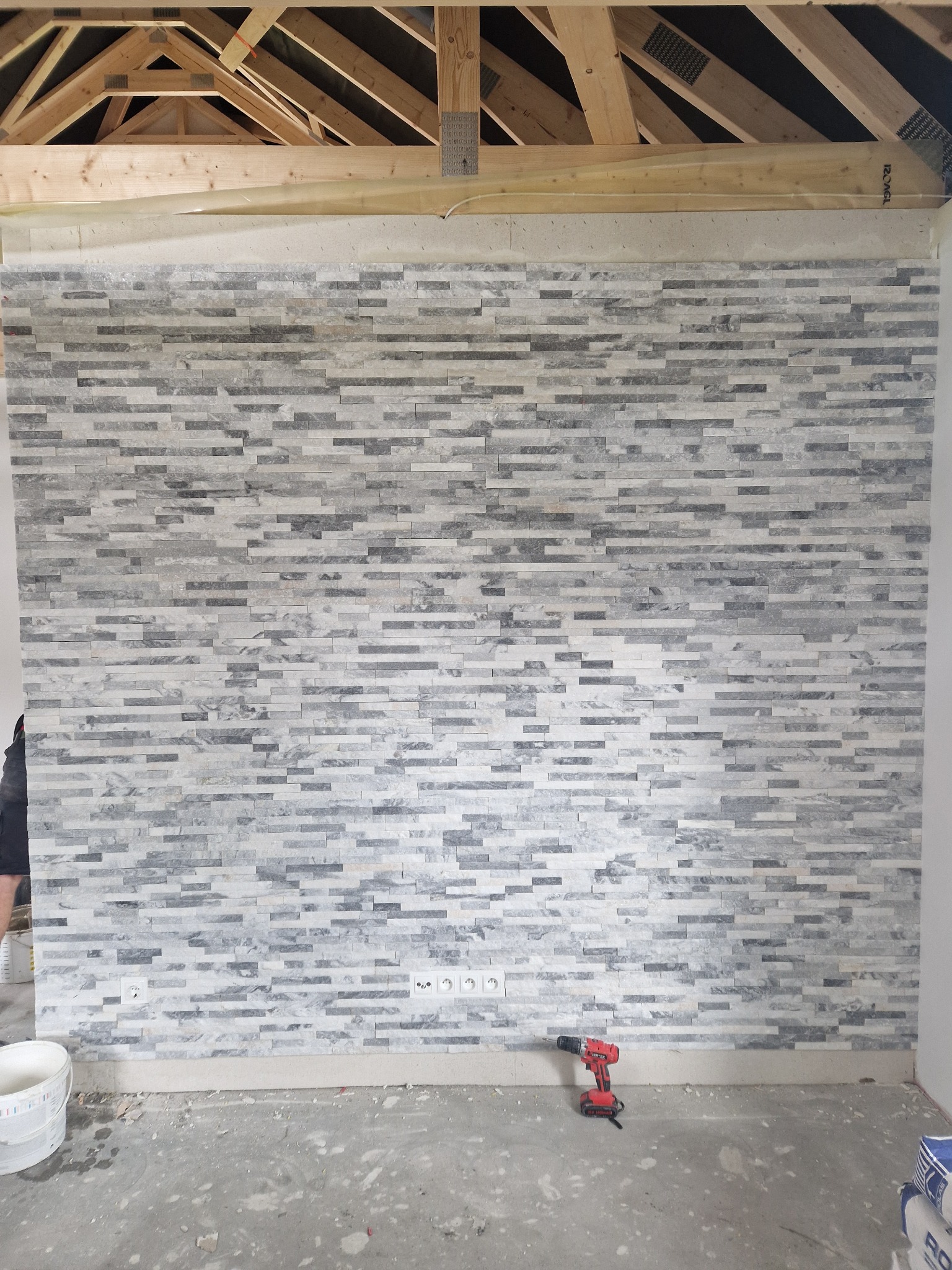 Ściana wyłożona dekoracyjną mozaiką z kamienia naturalnego w odcieniach szarości, widoczne gniazdka elektryczne, na podłodze wiertarka akumulatorowa i wiadro z farbą, surowy stan poddasza...