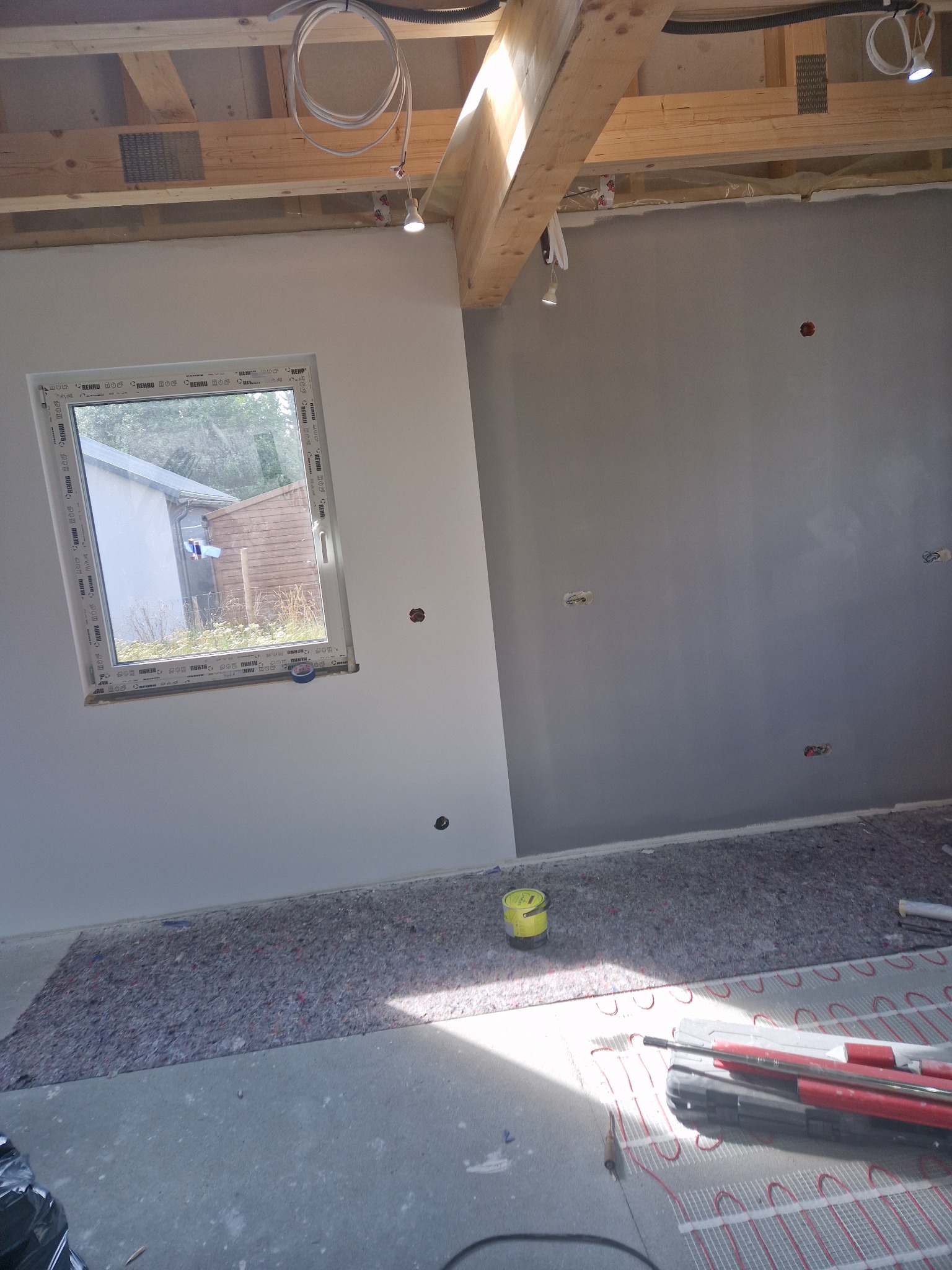 Wnętrze w trakcie prac wykończeniowych: częściowo pomalowana ściana na szaro, widoczne przygotowania do ogrzewania podłogowego, okno z widokiem na zewnątrz, żółte wiaderko i narzędzia na podłodze...