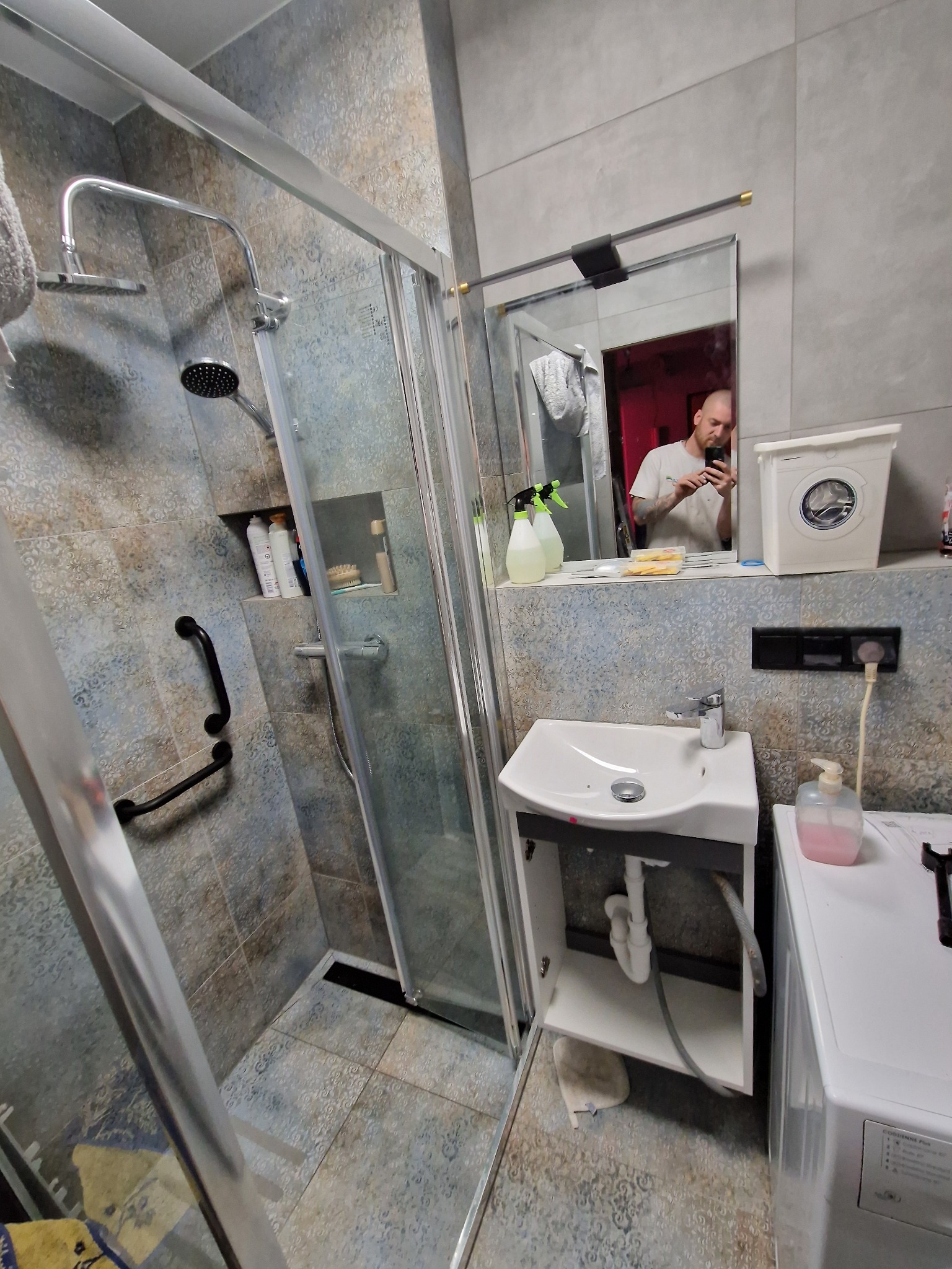 Umywalka z szafką, kabina prysznicowa z deszczownicą i pralka w łazience z szaro-beżowymi płytkami, widoczne akcesoria łazienkowe i odbicie fotografa w lustrze.