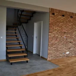 Wnętrze domu z drewnianymi schodami z czarną metalową balustradą, ceglaną ścianą z dwoma kinkietami i połączeniem szarych płytek z drewnianą podłogą.