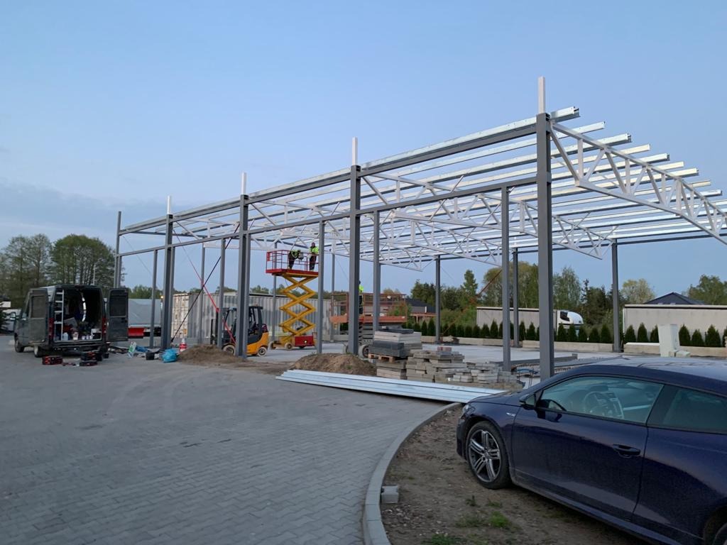 Montaż konstrukcji stalowej hali, widoczna podnośnik z pracownikami, elementy konstrukcyjne na ziemi oraz zaparkowane pojazdy