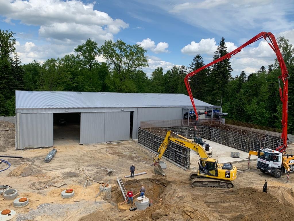 Budowa hali stalowej w trakcie: widoczny szkielet konstrukcji, betonowanie fundamentów, koparka Komatsu, pompa do betonu z wysięgnikiem, pracownicy w wykopie, elementy kanalizacji, las w tle...