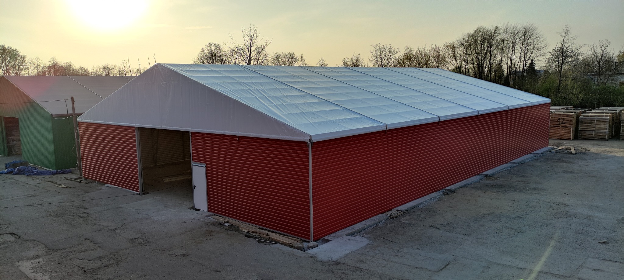 Czerwona hala magazynowa z białym dachem i drzwiami, widoczna z zewnątrz, z paletami składowanymi obok.