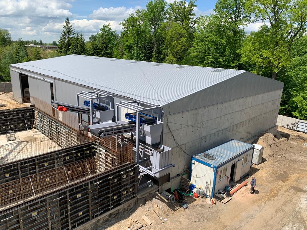 Wznoszenie hali produkcyjnej z betonu i stali, widoczne fundamenty i elementy konstrukcyjne, w tle zalesiony teren i niebieskie niebo.