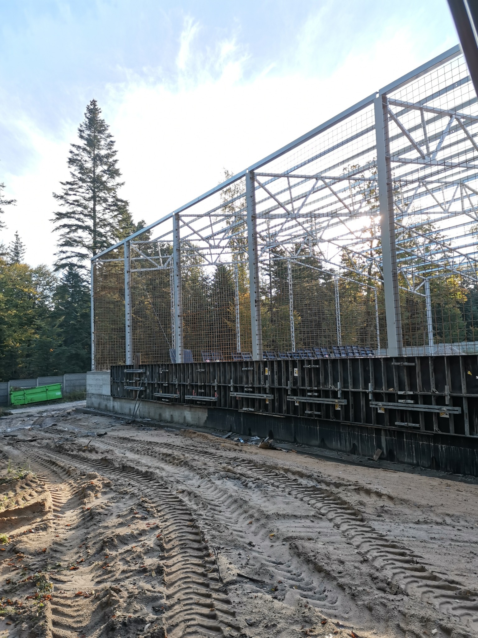 Konstrukcja stalowa hali w trakcie budowy, widoczne szalunki fundamentów, teren budowy z odciśniętymi śladami opon maszyn budowlanych, w tle las i błękitne niebo z promieniami słońca.