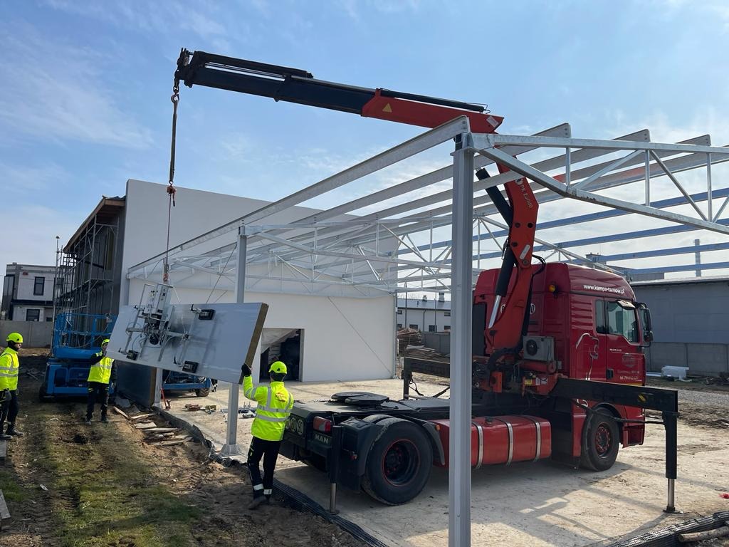Montaż płyty warstwowej na konstrukcji stalowej hali za pomocą przyssawki podwieszonej do dźwigu samochodowego, pracownicy w żółtych kamizelkach i kaskach, w tle budynek w trakcie budowy.