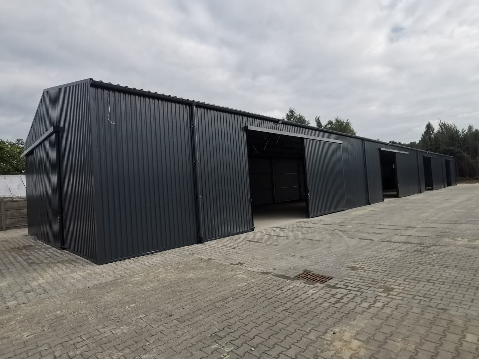 Szara hala magazynowa z blachy falistej z przesuwnymi drzwiami, widok z zewnątrz na betonowy plac, pochmurne niebo.