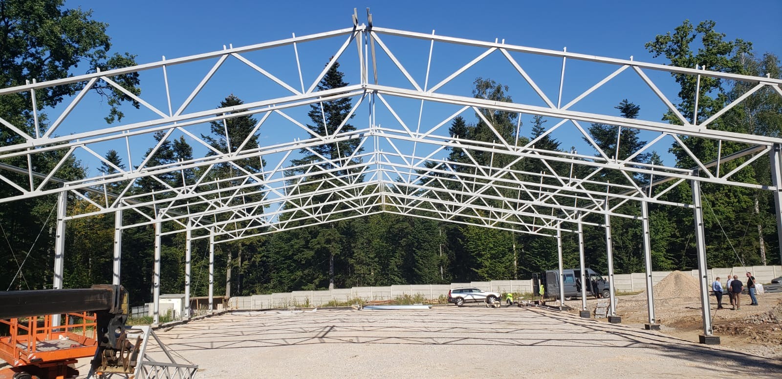 Konstrukcja stalowa hali w trakcie budowy, widoczne elementy nośne i kratownica dachu na tle lasu i błękitnego nieba, z pomarańczowym podnośnikiem koszowym po lewej stronie.