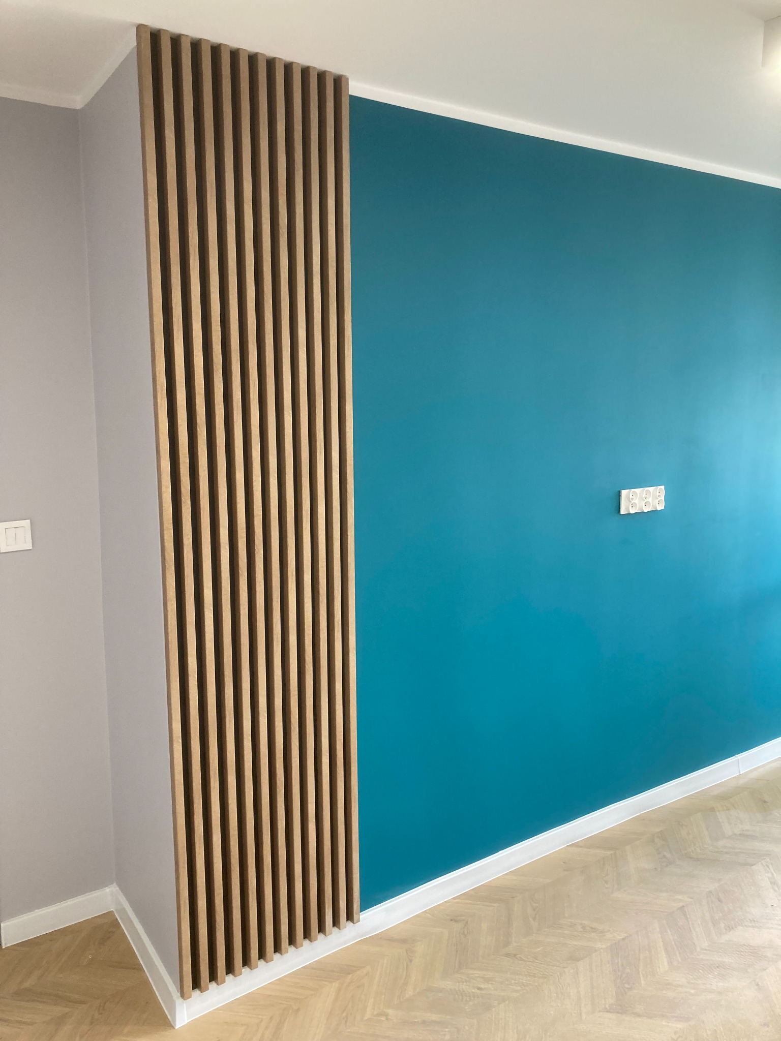 Nowoczesne wykończenie wnętrza z pionowymi lamelami drewnianymi na szarej ścianie, przylegające do ściany pomalowanej na turkusowo, z widocznymi gniazdkami elektrycznymi i parkietem w jodełkę...