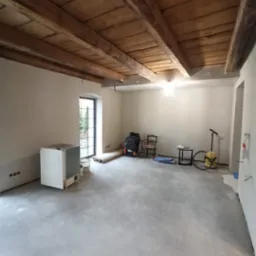 Surowe wnętrze w Brzegu przed wykończeniem, z drewnianym stropem i świeżo wylaną posadzką, widoczne narzędzia i sprzęt budowlany, przygotowane do dalszych prac.