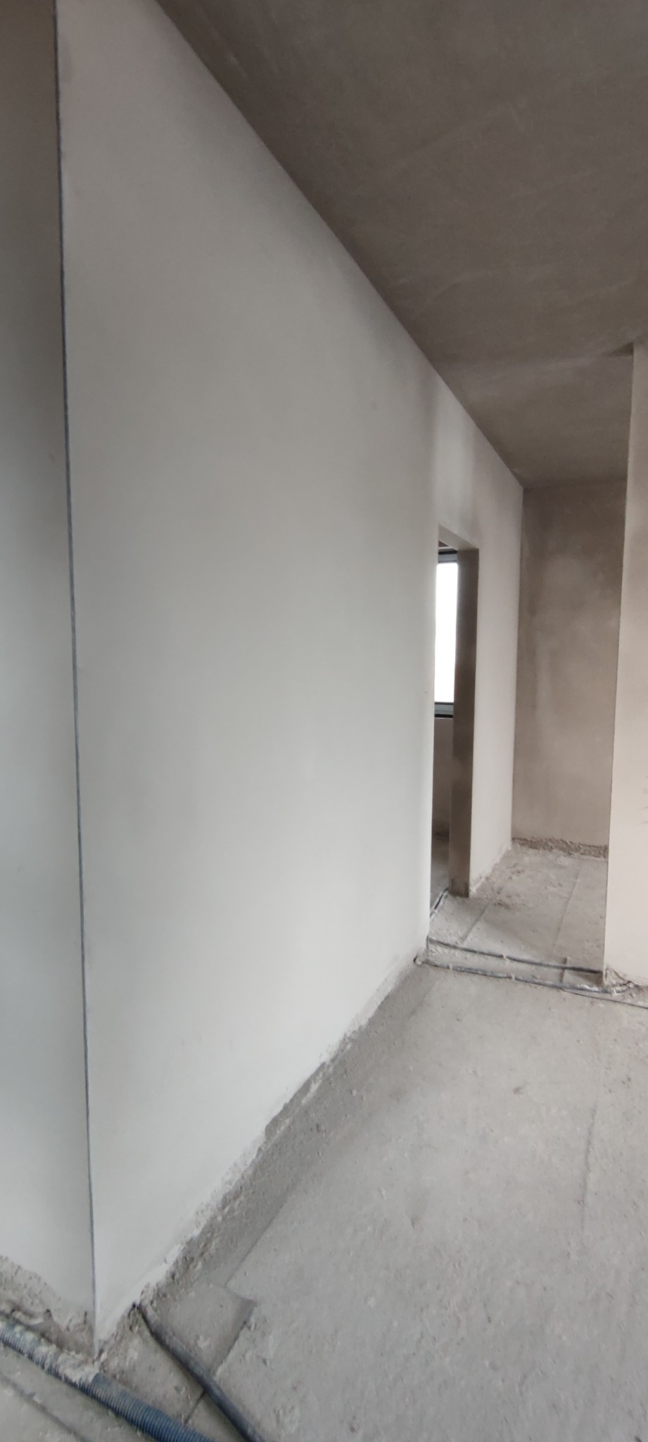 Świeżo otynkowane ściany w surowym stanie, widoczne przewody elektryczne przy podłodze, sufit w trakcie wykańczania.