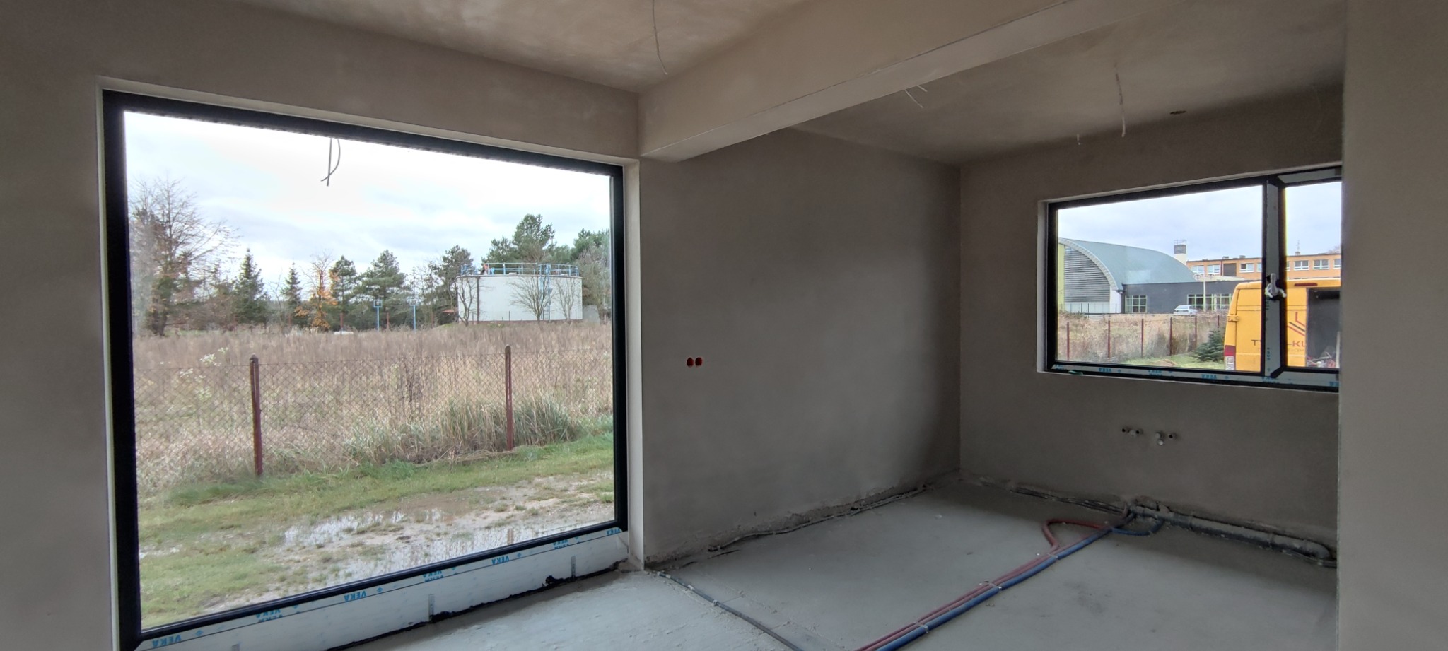 Surowe wnętrze po nałożeniu gładzi szpachlowej na ścianach i suficie, widoczne okna z czarnymi ramami, instalacja podłogowa i wystające kable ze ściany.