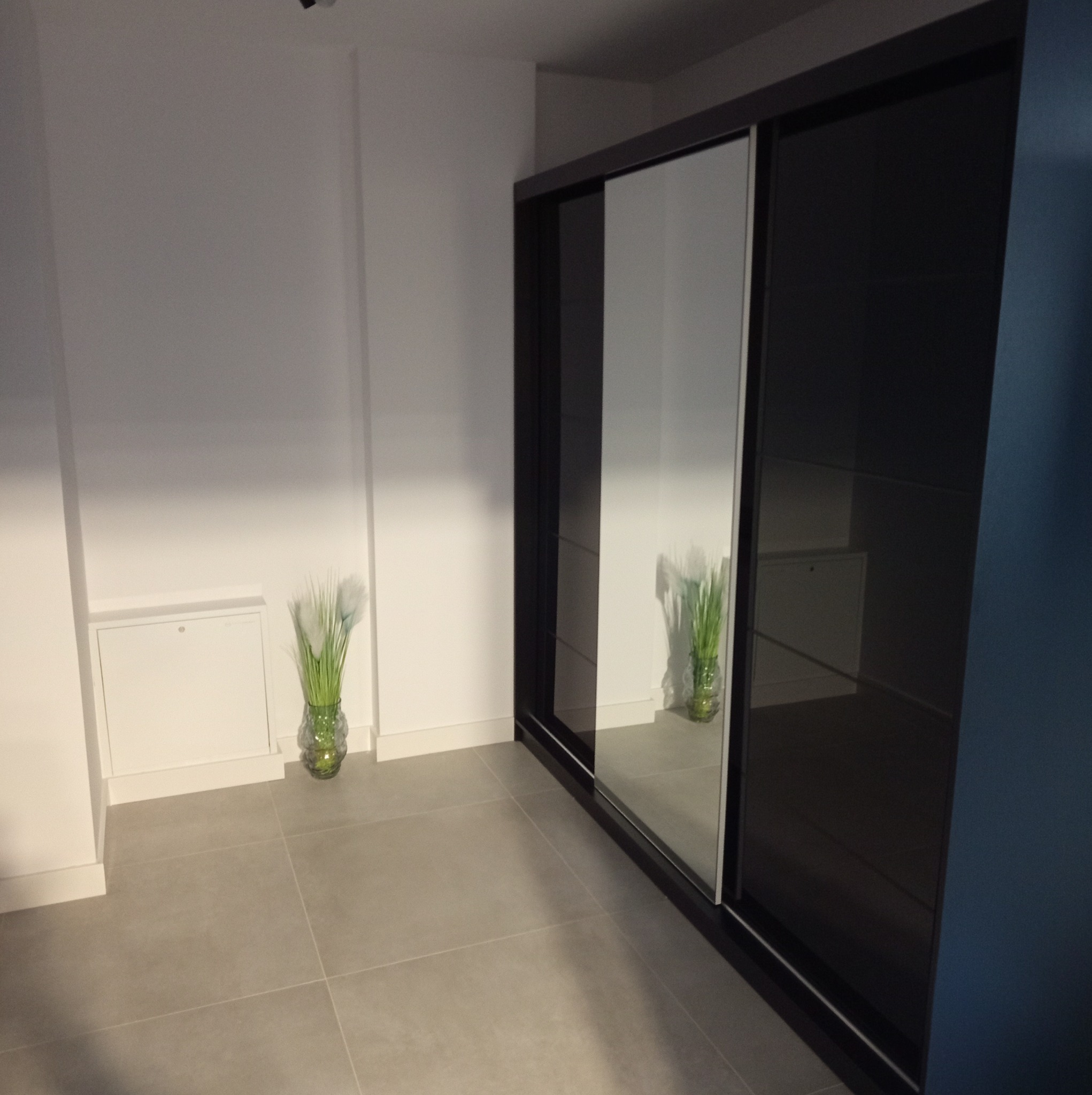 Nowoczesna szafa przesuwna z czarnymi ramami i lustrzanymi drzwiami w minimalistycznym wnętrzu z szarą podłogą i dekoracyjnymi trawami w szklanych wazonach.