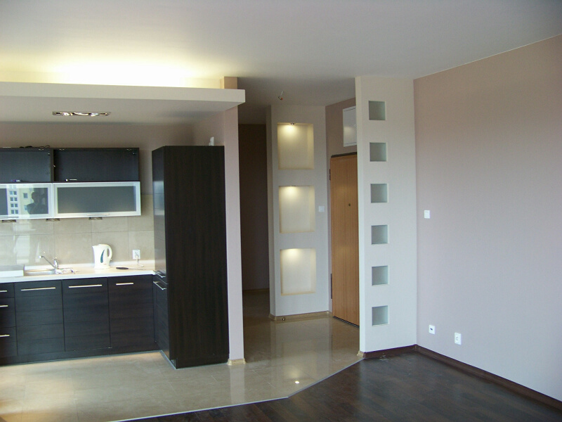 Nowoczesna kuchnia z ciemnymi szafkami i białym blatem, widok na przedpokój z dekoracyjną ścianką i podświetlanymi niszami.