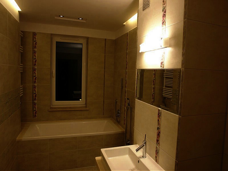 Wykończona łazienka z beżowymi płytkami, wanną pod oknem i nowoczesnym oświetleniem nad umywalką, z dekoracyjnym paskiem płytek z motywem kwiatowym.