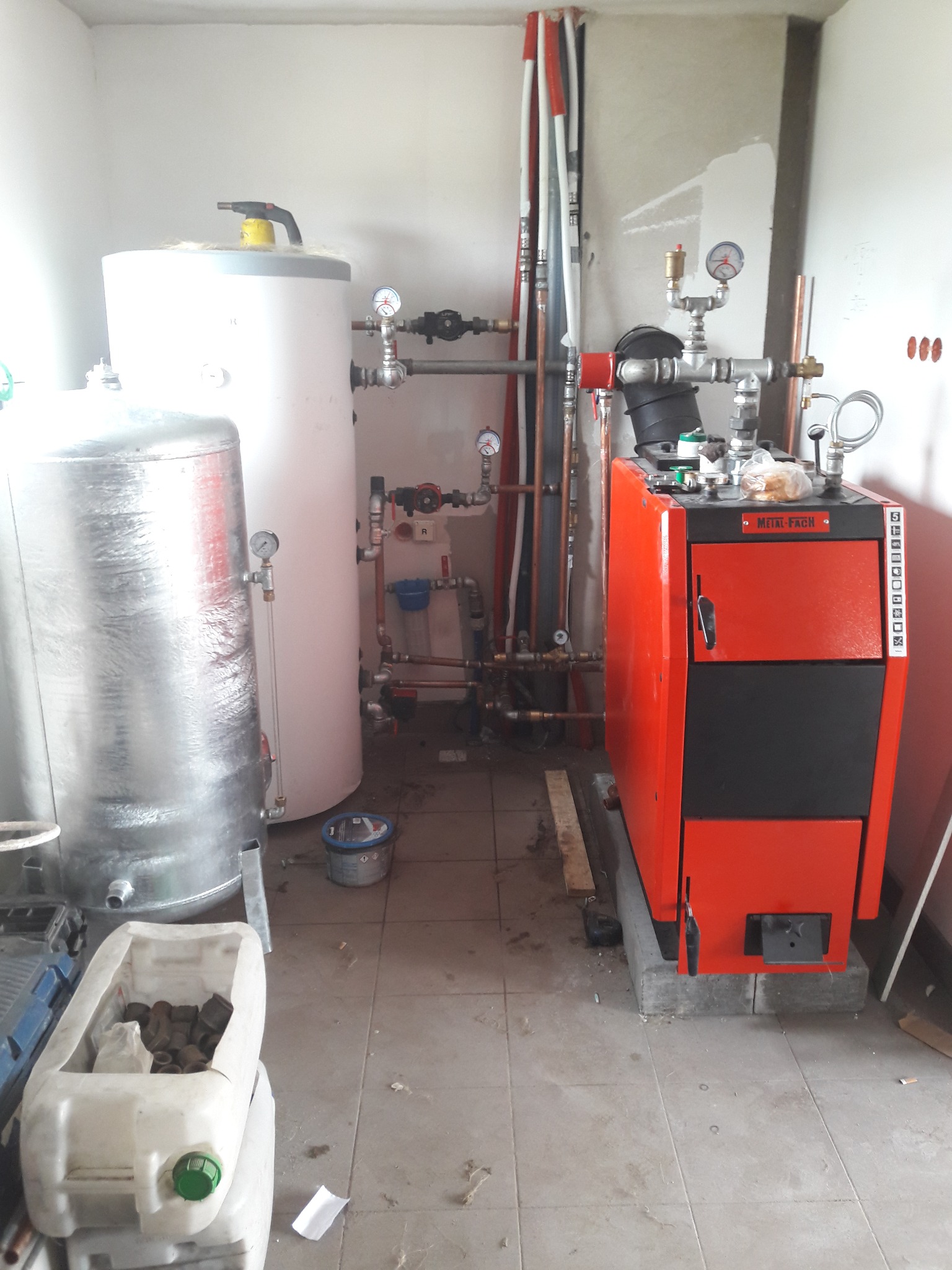 Kotłownia w trakcie montażu: czerwono-czarny piec Metal-Fach obok dwóch zbiorników, instalacja hydrauliczna z rurami, manometry, zawory i pojemnik z kształtkami na podłodze z płytek.