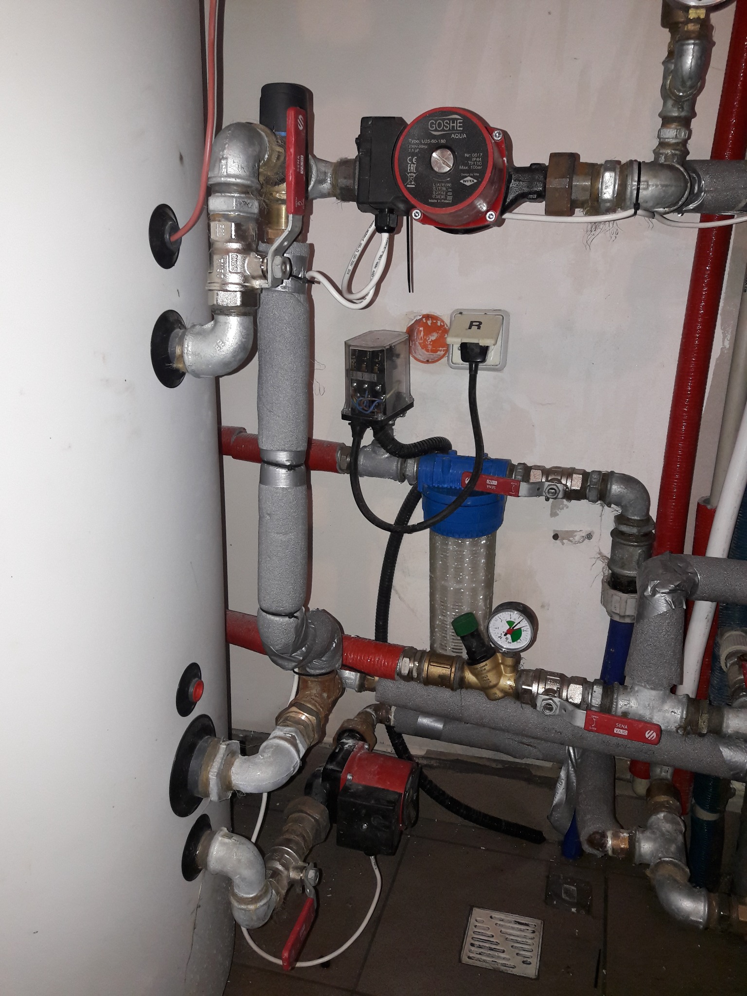 Instalacja centralnego ogrzewania z białym zasobnikiem, pompami obiegowymi Goshe i Sena, filtrem wody z manometrem, rurami stalowymi i miedzianymi, częściowo zaizolowanymi szarą pianką, widoczne...