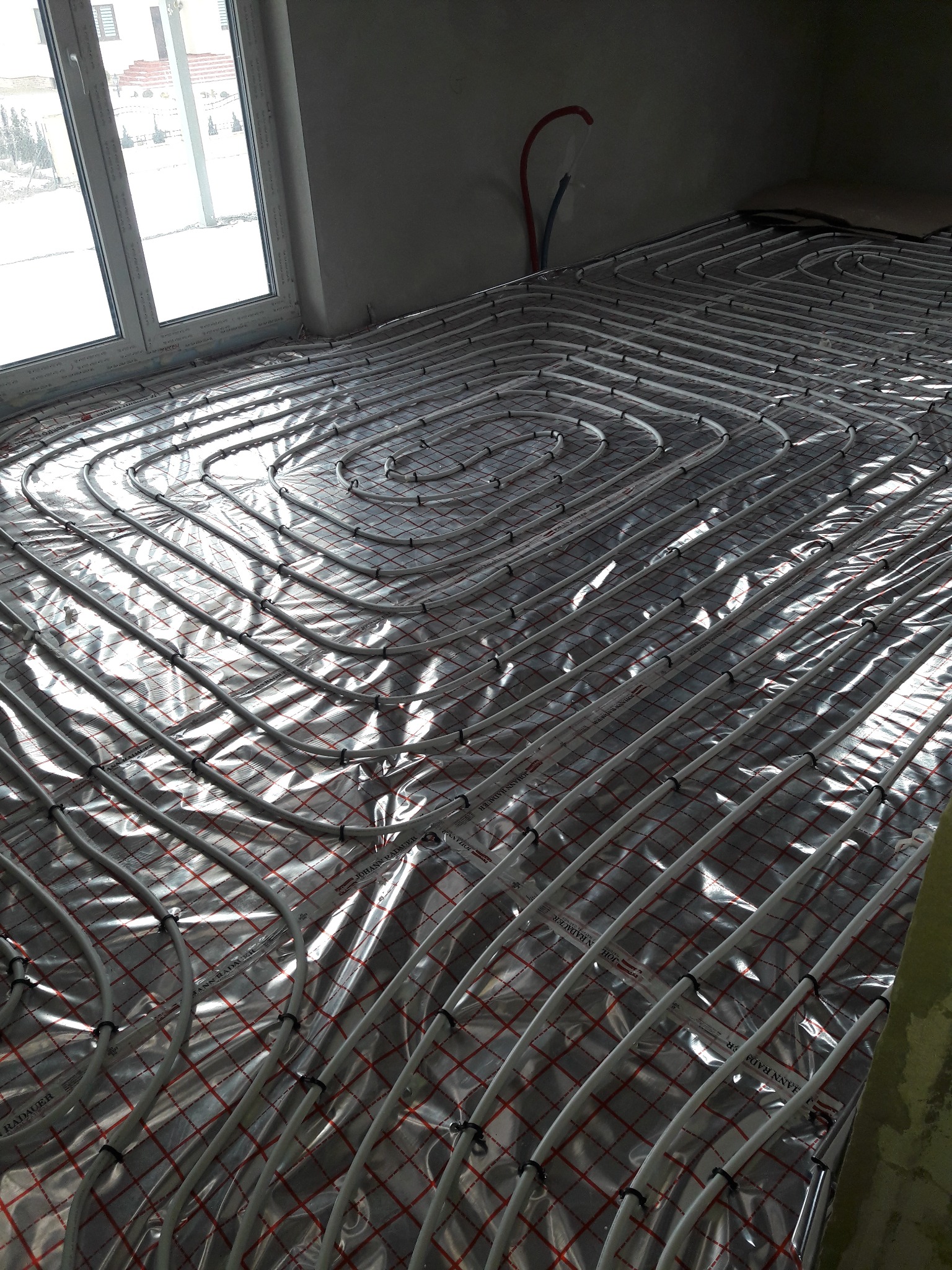Białe rury ogrzewania podłogowego, ułożone spiralnie na folii izolacyjnej z czerwoną kratką, w surowym wnętrzu z oknem.