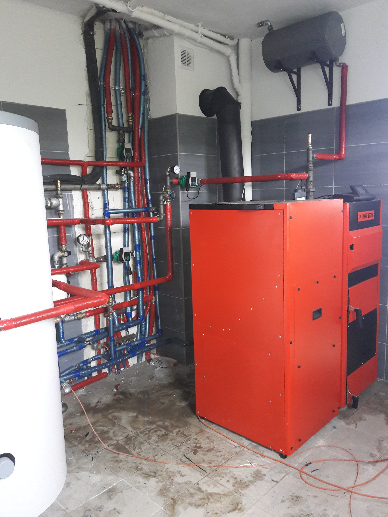 Czerwony piec CO z podłączonymi rurami w kolorach czerwonym i niebieskim, zbiornik wyrównawczy na ścianie, w tle szare płytki i biały zasobnik na wodę, widoczne przewody elektryczne na podłodze.